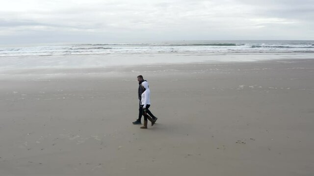 Couple walking on Oregon beach with white dog running, cloud cold day, orbit aerial view