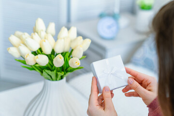 Woman holding small white gift box with jewelry and bouquet of white tulips flowers on woman day. Concept