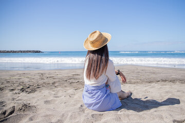 A girl wears a summer hat sitting on the beach and enjoys the sea. Vacation concept.