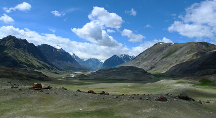 A wide alpine valley of the Akkol river in Altai with a small mountain in the middle in a mountain gorge with snow-capped peaks and mountain ranges against the background of a sky with clouds, summer