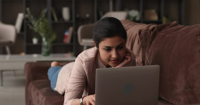 Relaxed millennial indian ethnicity woman lying on sofa, looking at laptop screen, thinking of creative business ideas, web surfing interesting information, enjoying spending leisure time online.