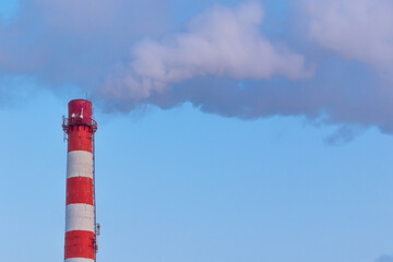 red and white chimney of a factory, from which gray smoke comes out against a blue sky on a sunny frosty winter day
