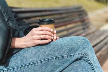 close-up take of the hands of a white woman holding a disposable paper cup of coffee. She wears blue jeans, black coat and a ring on her fingers. She is sitting on a wooden bench at an urban green