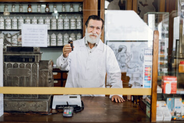 Portrait of handsome senior man pharmacist leaning on counter at old pharmacy and demonstrating bottle with medicine to camera.