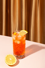 orange cocktail with lemon and ice on the table, gold fabric background, hard light, trendy still life