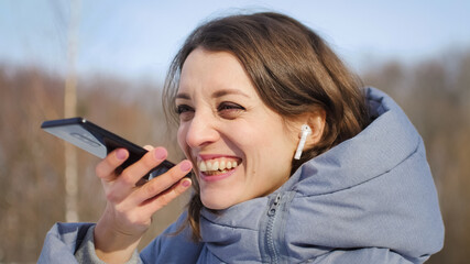 Young woman with wireless headphone is using her smartphone to send a voice message to a new social network Clubhouse standing somewhere outdoors at the park
