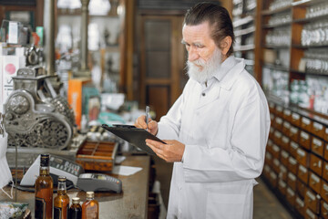 Fototapeta na wymiar Portrait of concentrated experienced male senior pharmacist writing on a clipboard while standing in beautiful vintage old pharmacy interior.