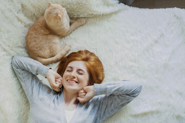 Smiling young red-haired woman lying on comfortable bed with ginger cat, stretching, waking up, enjoy good morning time