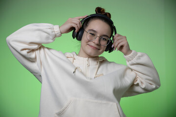 Smiling young teenage girl in modern wireless headphones having fun listen to music on green studio background