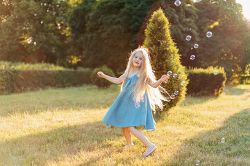 Child whirling, dancing plays on the meadow. Girl having fun with bubbles. Cute little longhair blonde girl dancing with soap bubbles at sunset park