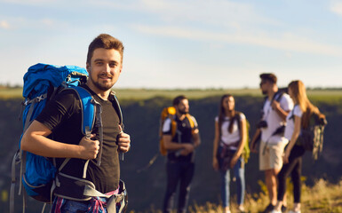 Fototapeta premium A young tourist man with a backpack looks at the camera on a hike against the background of friends hikers on nature in summer