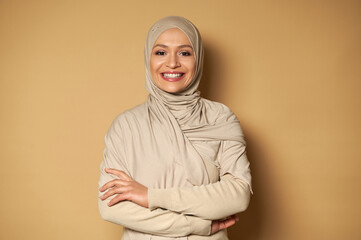 Smiling arabic Muslim woman posing with crossed arms on beige background with copy space