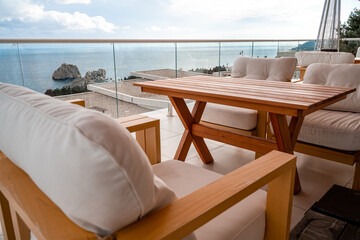Restaurant with sea view. You can see rocks in the sea. Wooden tables and chairs with white cushions. Overcast, cloudy weather.