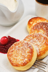 Cottage cheese pancakes or fried cottage cheese pancakes, sprinkled with powdered sugar, on a plate with cranberry jam and sour cream. A traditional Eastern European dish. Close-up, selective focus