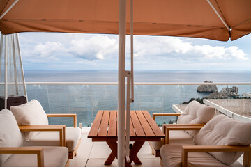 Obraz premium Restaurant with sea view. You can see rocks in the sea. Wooden tables and chairs with white cushions. Overcast, cloudy weather.
