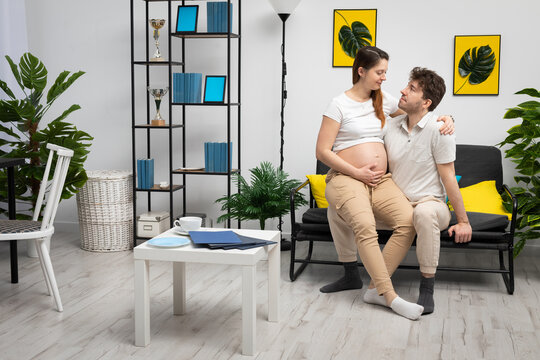 A pregnant wife is sitting on her husband's lap in a newly furnished room, holding her belly. A bright living room in a modern style with a preferable space.