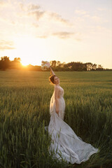 Fototapeta na wymiar Young happy bride in lace wedding dress holding bouquet of flowers in her hand and posing in the field in the summer sunset
