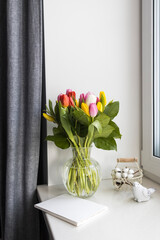 A bouquet of multi-colored tulips in a transparent vase on the windowsill on a holiday. White notebook, wicker metal basket with cotton flowers inside. Bird figurine.