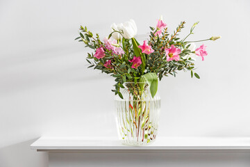 A minimalistic bouquet of white tulips, pink eustoma, hyacinth, eucalyptus in a fluted glass vase on a white panel of an artificial fireplace.