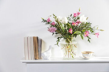 A minimalistic bouquet of white tulips, pink eustoma, hyacinth, eucalyptus in a fluted glass vase on a white panel of an artificial fireplace.