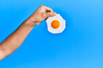 Hand of hispanic man holding fried egg over isolated blue background.