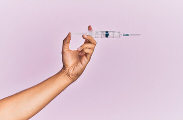 Hand of young hispanic man holding syringe over isolated pink background.