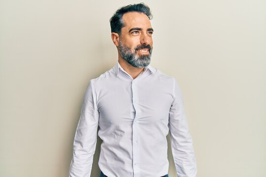 Middle age man with beard and grey hair wearing casual white shirt looking to side, relax profile pose with natural face and confident smile.