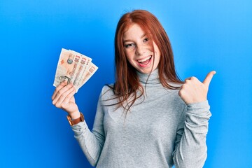 Young red head girl holding 10 united kingdom pounds banknotes pointing thumb up to the side smiling happy with open mouth