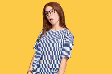 Young read head woman wearing casual clothes and glasses in shock face, looking skeptical and...