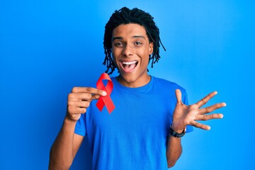 Young african american man holding support red ribbon celebrating achievement with happy smile and winner expression with raised hand