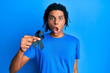 Young african american man holding black ribbon scared and amazed with open mouth for surprise, disbelief face