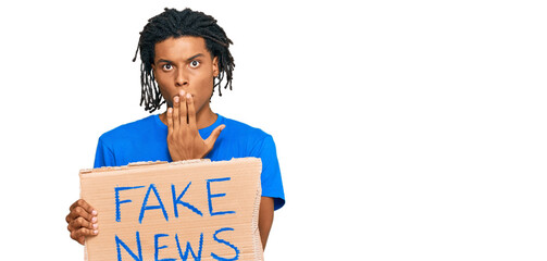 Young african american man holding fake news banner covering mouth with hand, shocked and afraid...