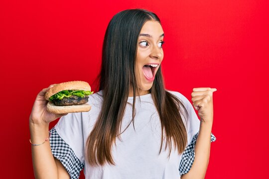 Young hispanic woman eating hamburger pointing thumb up to the side smiling happy with open mouth