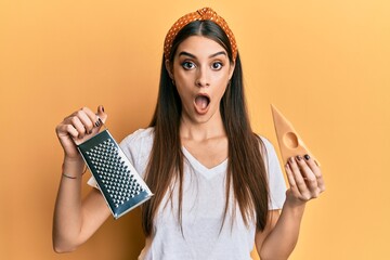 Beautiful brunette young woman holding grater and cheese afraid and shocked with surprise and...