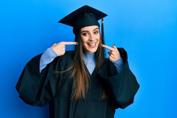 Beautiful brunette young woman wearing graduation cap and ceremony robe smiling cheerful showing...