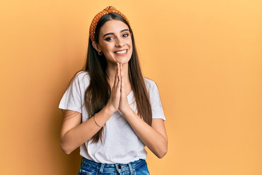Beautiful brunette young woman wearing casual white t shirt praying with hands together asking for forgiveness smiling confident.