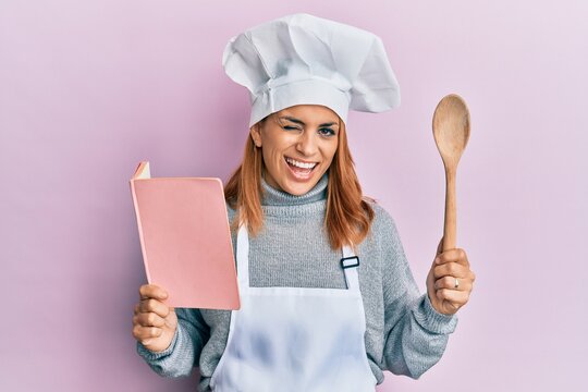 Hispanic young chef woman reading recipes book winking looking at the camera with sexy expression, cheerful and happy face.