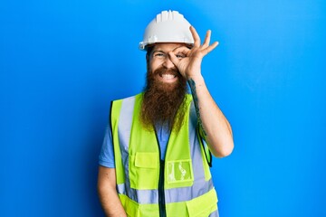 Redhead man with long beard wearing safety helmet and reflective jacket doing ok gesture with hand smiling, eye looking through fingers with happy face.