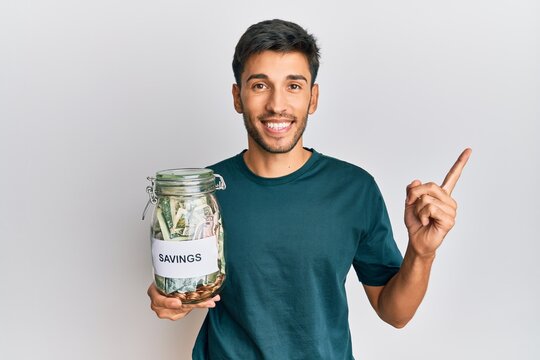 Young handsome man holding jar with savings smiling happy pointing with hand and finger to the side