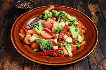Frozen cut Vegetables, broccoli, sweet peppers, tomatoes, carrots, peas and corn on a plate. Dark Wooden background. top view