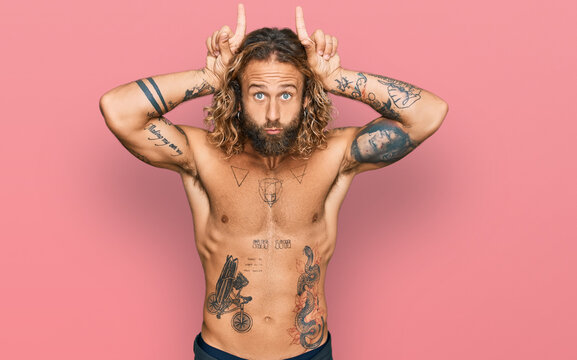 Handsome man with beard and long hair standing shirtless showing tattoos doing funny gesture with finger over head as bull horns