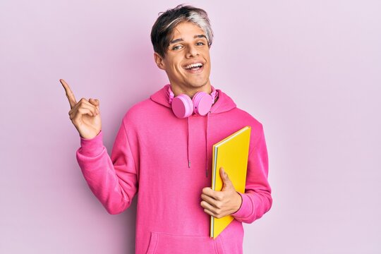 Young hispanic man holding book using headphones smiling happy pointing with hand and finger to the side