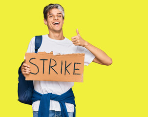 Young hispanic man holding strike banner cardboard smiling happy and positive, thumb up doing excellent and approval sign