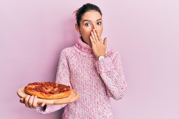 Young caucasian woman holding italian pizza covering mouth with hand, shocked and afraid for...
