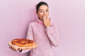 Young caucasian woman holding italian pizza covering mouth with hand, shocked and afraid for mistake. surprised expression