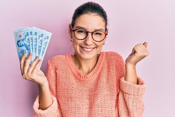 Young caucasian woman holding 50 thai baht banknotes screaming proud, celebrating victory and success very excited with raised arm