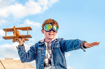 Fashionable teenager boy in pilot glasses with an wooden airplane in his hand against the blue sky...