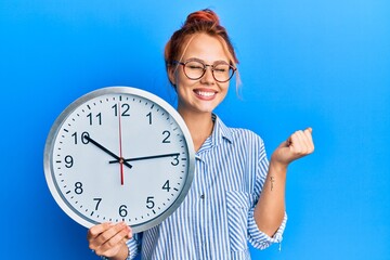 Young redhead woman holding big clock screaming proud, celebrating victory and success very excited...