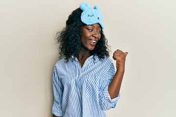 Beautiful african young woman wearing sleep mask and pajama smiling with happy face looking and...