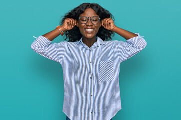 Beautiful african young woman wearing casual clothes and glasses smiling pulling ears with fingers,...